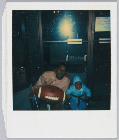 [Man holding large football posing next to baby in stroller, large camera flash glare behind them]