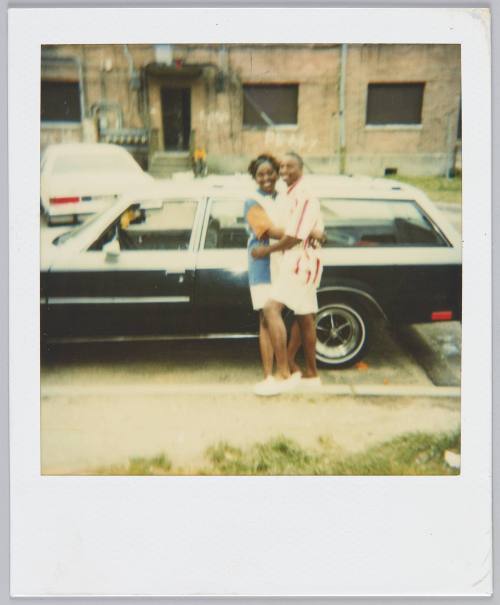 [Young couple hugging in front parked car, man on right wears red and white striped shirt]