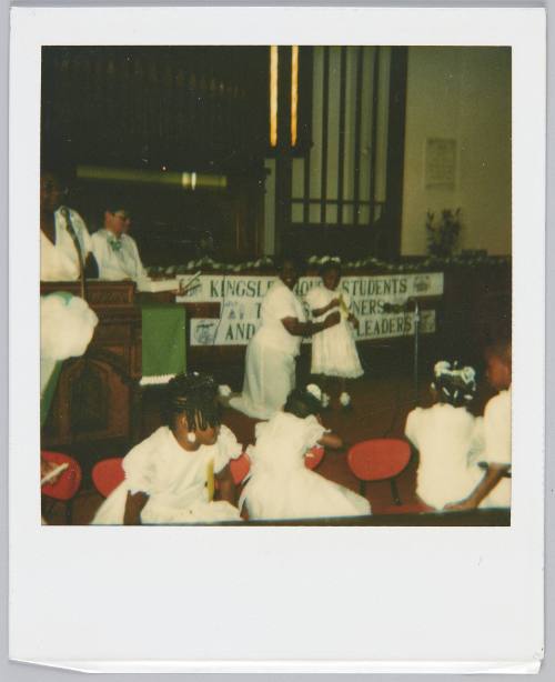 [Little girl in white dress standing in front of school group in matching dresses, woman in white kneels beside her]