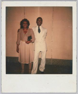 [Man in all white suit with arm around older woman in tan outfit]