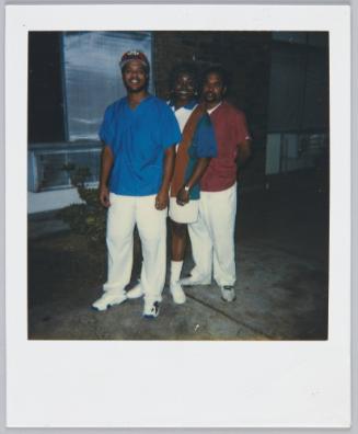 [Woman standing in between two men, man on left wearing blue shirt and white pants]