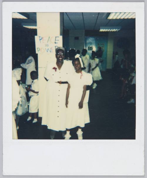 [Elderly woman and girl in white dresses standing in front of pillar with sign that reads 'Peace is Power']
