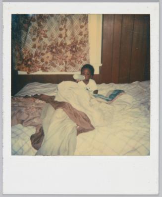 [Baby drinking from bottle sitting in the middle of a bed]