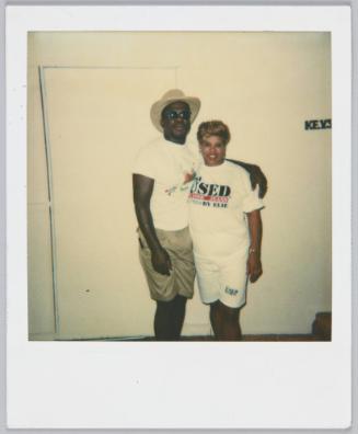 [Man in graphi tshirt and khaki shorts with arm around woman in white graphic tshirt and white long shorts]