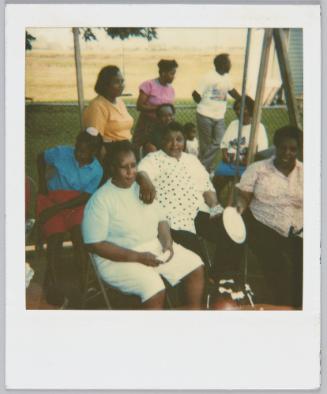 [Family sitting around on lawn chair, woman on right holding out paper plate]