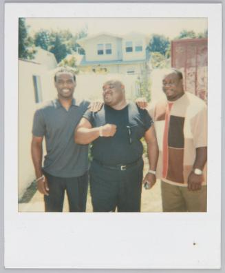 [Group of three men standing outside, man in the middle wearing all black with one hand placed on his chest]