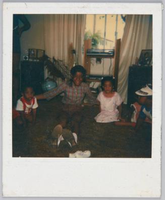 [Little girl in plaid sitting on rug in between toddler boy and girl]