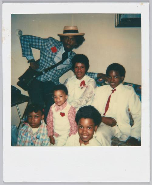 [Group in formal attired gathered on and around couch, man in back wears a blue plaid blazer]