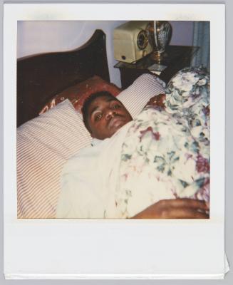 [Close up of man with moustache laying in bed with covers pulled up to his chin]