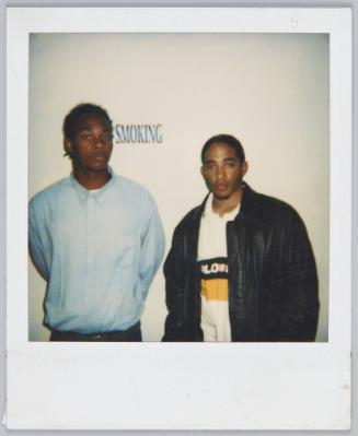 [Two young men standing in front of white wall, 'SMOKING' sign on wall behind them]