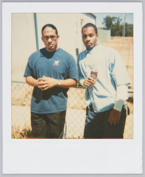 [Two young men leaning by metal fence, man on right holding a can of soda]