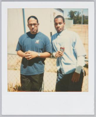 [Two young men leaning by metal fence, man on right holding a can of soda]