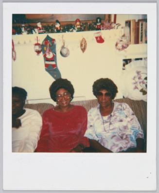 Dec. 7, 1985 [Two women wearing big glasses sitting on couch, wall with Christmas decorations behind him]