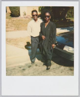 [Couple standing in street in front of car, both wearing black sunglasses]