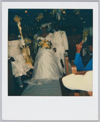 [Newlywed couple walking down the aisle, bride wears a white hat and holds bouquet of white flowers]