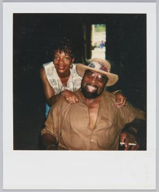 [Man in brown short sleeve button up and hat smiling, woman leaning on his shoulders behind him]