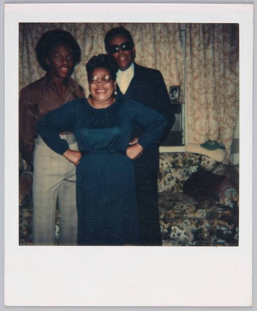 [Woman wearing black dress and glasses standing in between two men, man on left side has an afro]