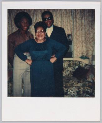 [Woman wearing black dress and glasses standing in between two men, man on left side has an afro]