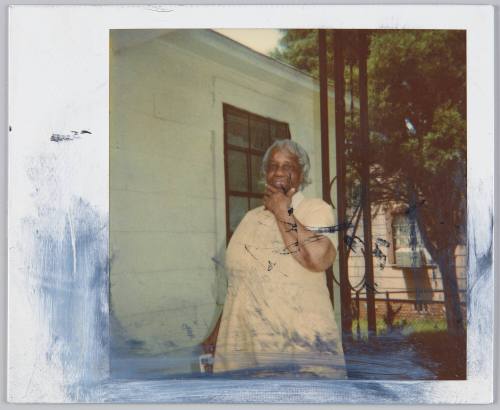 [Elderly woman standing on porch with hand rested on chin]