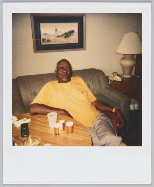 [Elderly man in yellow tshirt sitting on couch, painting of a lighthouse on the wall behind him]