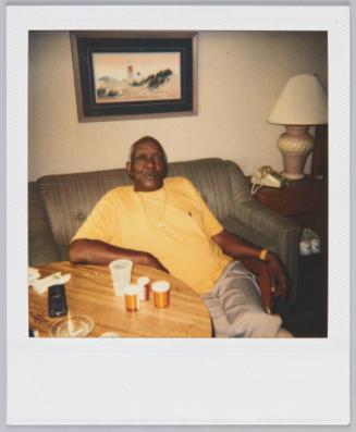 [Elderly man in yellow tshirt sitting on couch, painting of a lighthouse on the wall behind him]