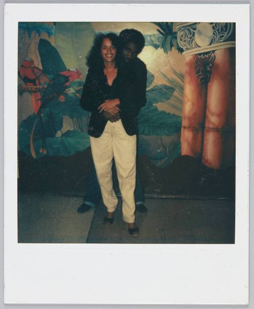 [Couple in front of decorated wall, man standing behind woman with his arms around her]