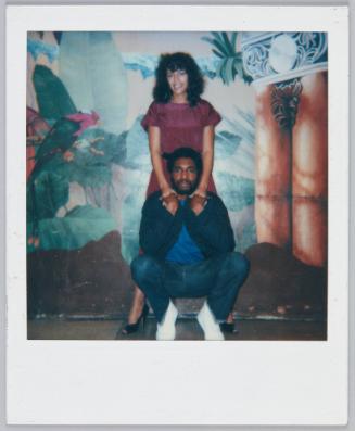 [Couple in front of decorated wall, man squatting and woman stands behind him as they hold hands]