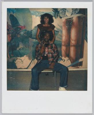 [Couple in front of decorated wall, man sits on chair and woman stands behind him]