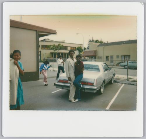 [Couple arm-in-arm posing in parking lot, in front of white car]