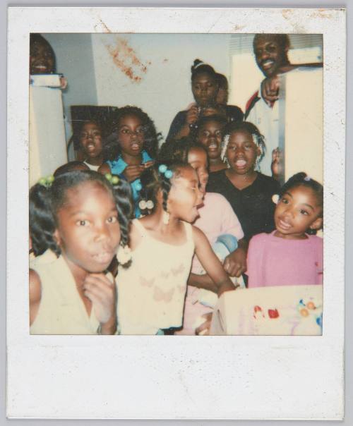 [Eight girls standing in group wearing colourful hair accessories, adults smiling in background]