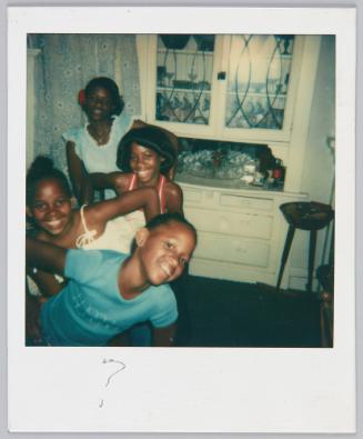 [Four girls standing in a line looking over each others shoulders and smiling with hands on hips in room with white cabinet]