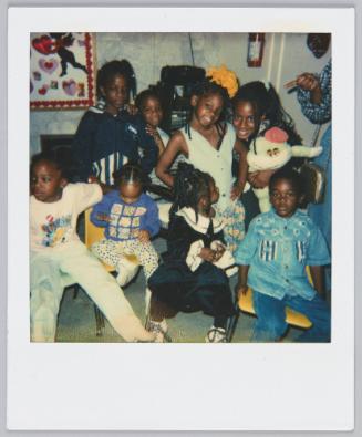 [Four children sitting on yellow chairs, four girls gathered around behind the seated children, girl standing on far right is holding a frog plush toy]