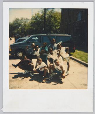 [Nine children crouching in a group in parking lot in front of large van]