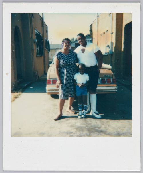 6-16-85 [Man, woman and child standing in driveway in front of red car between two houses]