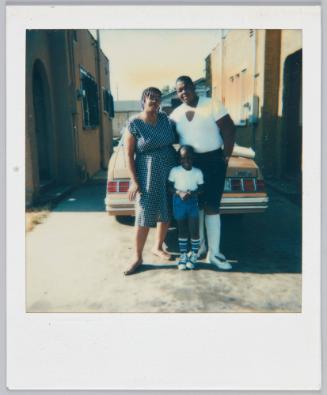 6-16-85 [Man, woman and child standing in driveway in front of red car between two houses]