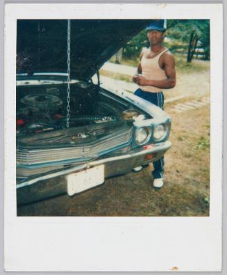 [Man in tank top and sweatpants standing next to car with the hood propped open]