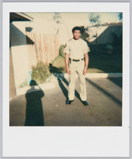 [Man in uniform standing in driveway, shadow of photographer can be seen on left side]