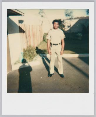 [Man in uniform standing in driveway, shadow of photographer can be seen on left side]