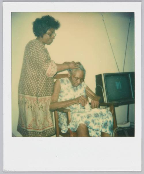 [Woman touching elderly woman's head while she sits in wooden chair beside television set]
