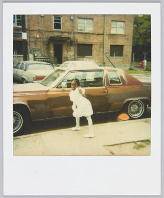[Girl in white dress standing on sidewalk in front of brown car making a peace sign with her tongue sticking out of her mouth]