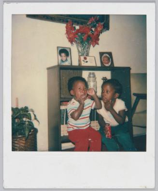 [Child crying with hands clasped on face sitting on shelve with another child]