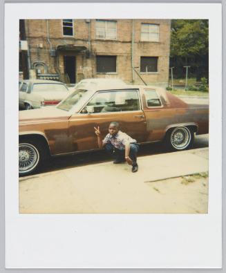 [Child crouching next to brown car on street making a peace sign]