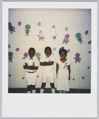 [Two boys and a girl in white outfits wearing blue ribbons on the left side of their chest]