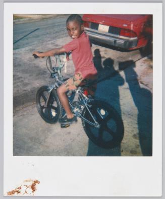 [Boy on bicycle on sidewalk next to red car, shadow of photographer seen on the right]