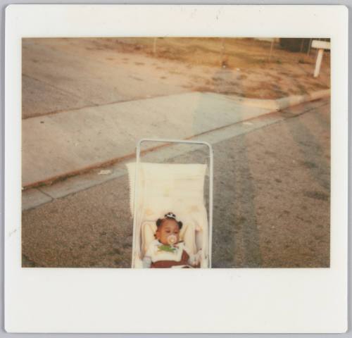 [Baby in carriage with soother in her mouth on street, shadows of people on the right side]