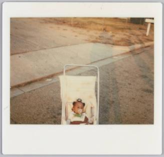 [Baby in carriage with soother in her mouth on street, shadows of people on the right side]