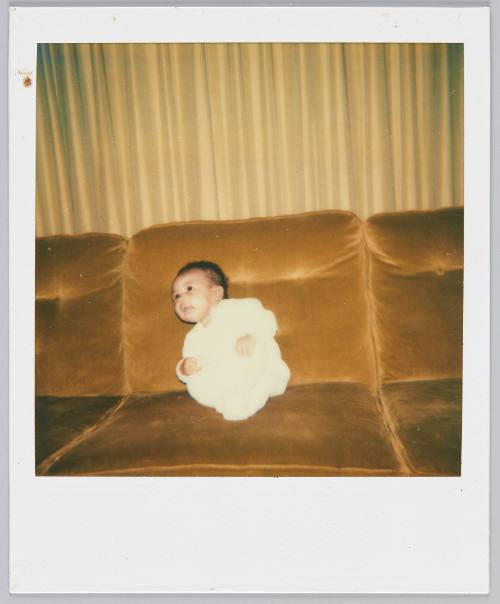 [Baby in white sitting on velvet couch looking up to the left]