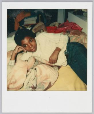 [Boy laying on his side on bed propping his head up with his arm, red plastic bag behind him]