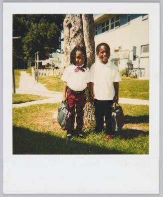 [Little boy and girl smiling and standing together with school bags in front of tree]