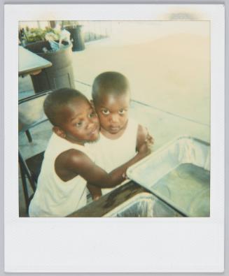[Two boys hugging wearing matching white tank tops standing beside table with aluminum trays]